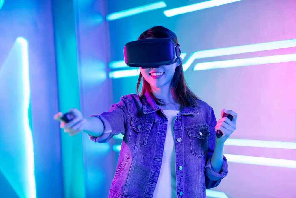 Person with long brown hair wears a VR headset, playing a game with hand held nunchucks. They wear a denim jacket and stand against a neon blue and pink background.